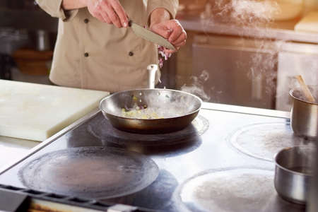Chef cutting onion into frying pan. Male chef at professional kitchen cutting onion in frying pan with stewed vegetables. Culinary and cuisine concept.の写真素材