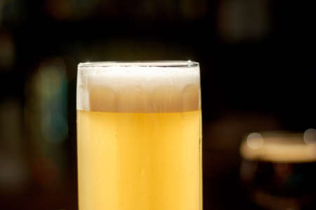 Cold lager beer on counter. Glass of fresh beverage with foam on dark bar background. Cropped image...