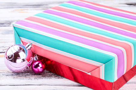 Colorful gift box and Christmas balls. Multicolored striped gift box and Christmas baubles on wooden background close up. Merry Christmas and happy New Year.の写真素材