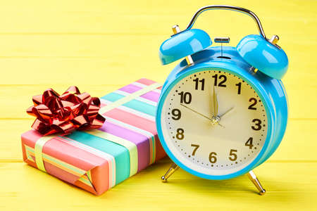 Gift box with bow and alarm clock. Christmas striped gift box with red bow and blue classic alarm clock on yellow wooden background. Time for New Year greeting.の写真素材