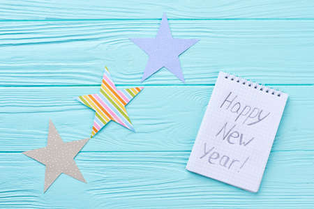 Cut out paper stars and paper notebook. Handmade paper stars and paper notepad with text Happy New Year on blue wooden background, top view. New Year wooden background.の写真素材