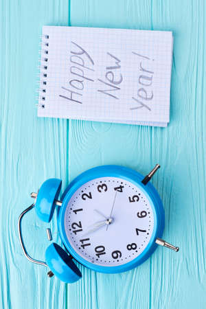 New Year decoration on wooden background. Blue alarm clock and paper notebook with message Happy New Year on blue wooden background, top view.の写真素材