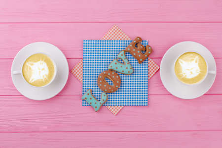 Word love from baked cookies. Couple of cappuccino cups with cats and cookies on pink wooden background. Valentines Day morning breakfast.の写真素材
