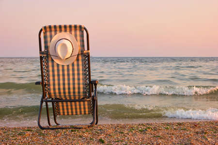 Rear view beach recliner in sea wave background. Deckchair with attached sun hat on the back.