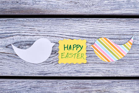 Easter yellow card and papercut figurines. Happy Easter inscription and cut out paper birds on old wooden background. Kids handmade paper ornaments for Easter holidays.の写真素材