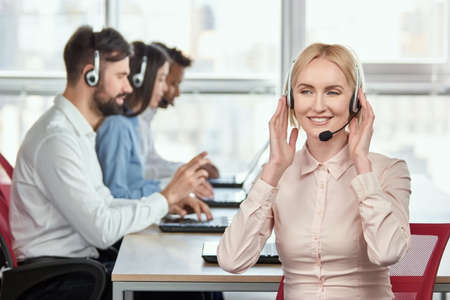 Call center service operator listening to headset. Smiling mature blond lady listening to client...