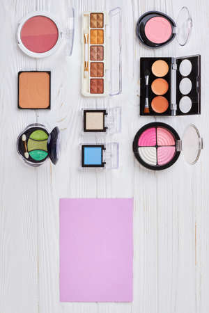 Makeup products kit, top view. Set of multicolored eyeshadows next to blank paper sheet on white wooden background. Fashion makeup cosmetics.の写真素材