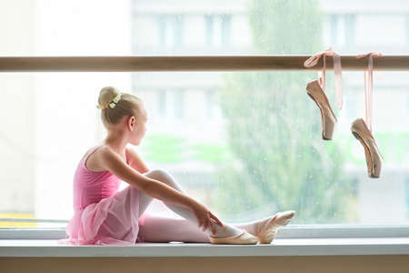 Ballerina in pink dress sitting on window-sill. Young beautiful ballet girl looking at the window. Pair of pointe shoes hanging on ballet barre.の写真素材