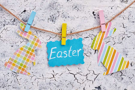 Easter card and papercut patterned rabbits. Cut out paper bunnies and blue paper with text Easter hanging on rope with clothespins on grey textured background. Easter decoration at home.の写真素材