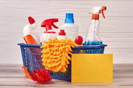 House cleaning products in basket. Bottles with cleaning liquid, brush, microfiber glove and blank...