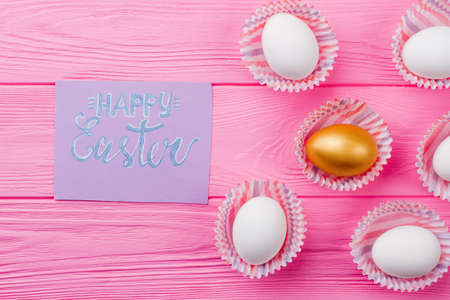Happy Easter composition with eggs. White eggs and one gold egg in paper wrappers on pink wooden table. Happy Easter concept.の写真素材