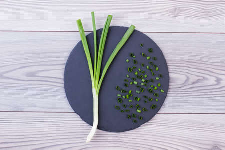 Whole and chopped green onion, top view. Fresh spring onion on round slate. Young onion against grey...