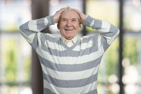 Shocked senior man touching his head. Portrait of old grandfather in windows background.