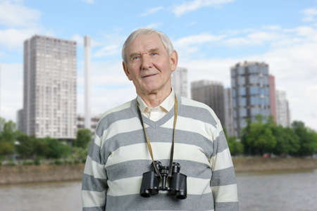 Senior with binoculars round his neck. Portrait of old man. City river background.の写真素材