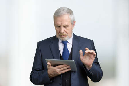 Old businessman with tablet. Portrait of pensive businessman with modern device. Bright blurred background.の写真素材