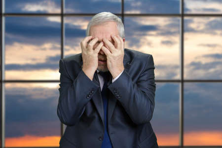 Portrait of an old stressed business man. Senior man in suit extremely sad and dissapointed, blue evening sky background with checkered windows.の写真素材
