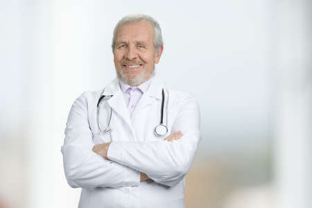 Portrait of doctor standing with arms crossed in hospital. Wrinkled caucasian physician. Bright blurred background.の写真素材