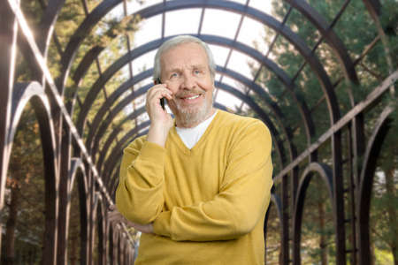 Smiling old grandfather talking on phone in greenhouse. Portrait of cheerful senior man in metal outdoor framework background.の写真素材