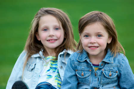 Close up portrait of two little sisters. Caucasian kids with denim jackets on the grass.