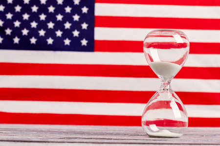 Glass timer with USA flag in the background. Hourglass on wooden table with flag of United States of the America in the background, horizontal image.の写真素材