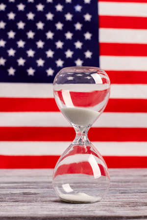 Hourglass on table with USA flag in the background. Sand glass on old wooden table with American flag in the background, vertical image.の写真素材