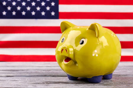 Money box on USA flag background. Yellow piggy bank on old wooden table with blurred American flag in the background. Financial and economics concept.の写真素材