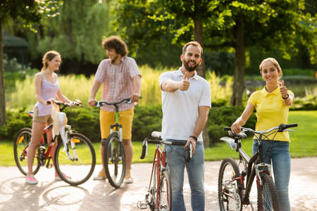 Young cheerful couple with bicycles. Happy smiling man and woman gesturing thumbs up on summer park background. Enjoying the summer bike ride.の写真素材