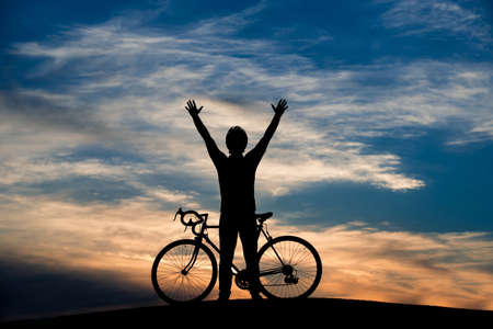 Young cyclist raised hands at sunset sky. Enjoying beauty of evening sky. Active summer rest and healthy lifestyle. Freedom and life energy.の写真素材