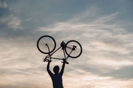 Silhouette of man holding bike above head. Man with bike above head on evening sky background. Concept of winner.の写真素材