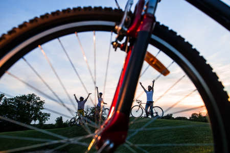 Young happy cyclists outdoors. Friends with bikes on green hill on evening sky background. Wheel of bicycle close up.の写真素材