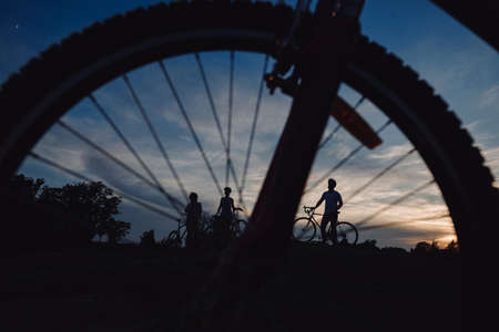 Cyclists through bicycle wheel. Silhouettes of cyclists on evening sky background.の写真素材