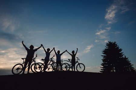 Group of cyclists holding hands together at sunset. Silhouettes of young persons with bikes standing on meadow and holding hands at evening sky.の写真素材