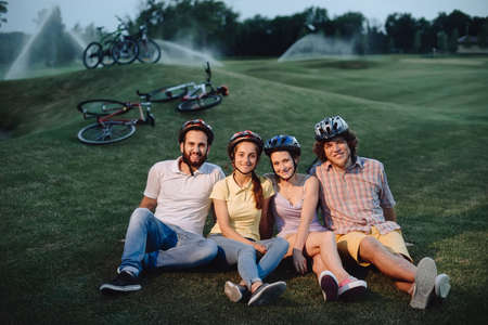 Four friends resting on green meadow. Group of cute cyclists sitting on the grass and looking at camera. Tourist are resting in park.の写真素材