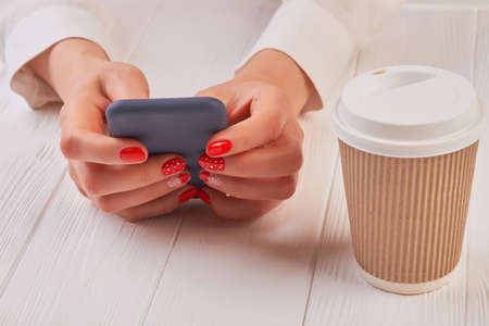 Female hands with smartphone and coffee. Woman manicured hands holding touch screen phone next to cardboard disposable cup with cappuccino.の写真素材