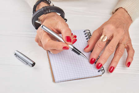 Old woman manicured hands with pen. Aged female hands with with pen and paper notebook on white wooden background.の写真素材