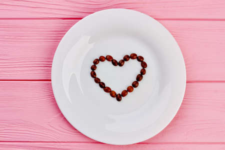 Heart from coffee beans on white plate. White plate with heart shaped roasted beans, top view....