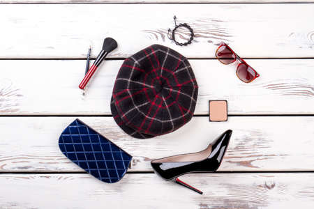 Woolen checkered hat and cosmetic makeup accessories, flat lay. Blue wallet, shiny high heel shoe, spectacles and brush. White wooden background.の写真素材