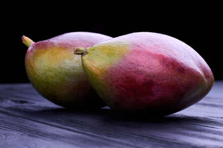 Close up fresh whole mangoes. Two tropical mango fruits on black wooden surface. Mango nutrition facts. Tasty exotic fruit.の写真素材