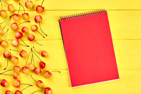 Yellow background with ripe cherries and notebook. Red and yellow cherries with red closed notepad on color background. Note the calories.の写真素材