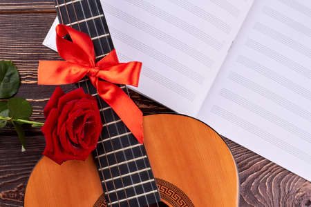 Guitar and musical notes on wooden background. Retro guitar with red ribbon. Rose and sheets of musical notes. Romantic musical background.の写真素材
