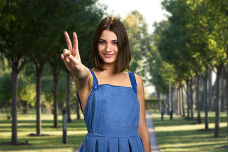 Beautiful girl with victory sign outdoor. Pretty teenage woman showing sign of peace with two fingers on summer nature background.の写真素材