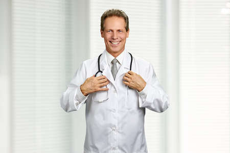 Joyful male doctor with stethoscope. Middle-aged practitioner with medical equipment is smiling on hospital background. Physician at work.の写真素材