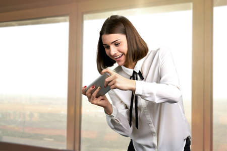 Funny girl playing game on smartphone. Happy cheerful young brunette playing video game on her phone on window background.の写真素材