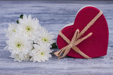 Red heart-shaped gift box and white flowers. Bunch of chrysanthemum flowers and red box in a shape of heart on wooden background. Happy Valentines Day.の写真素材