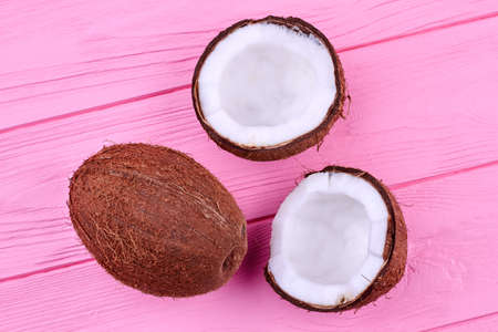 One whole and two cracked coconut halves on pink wooden background.