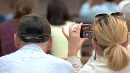 Woman taking photo of music concert. Mature couple at concert outdoor. Woman using smartphone, back...