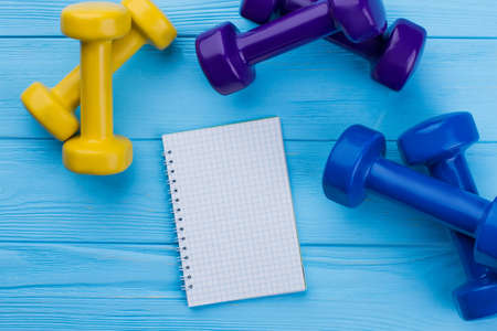 Three pairs of dumbbells and blank notebook. Colorful dumbbell weights and empty paper notepad on color background. Women items for sports life.の写真素材