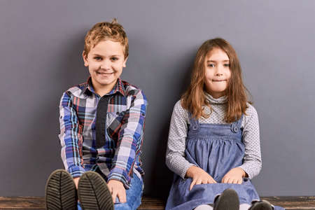 Little cute sister and brother portrait. Adorable pretty kids sitting near wall and smiling. Studio shot of positive happy little brother and sister.の写真素材