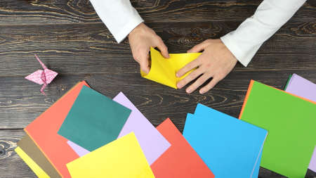 Male hands making origami with yellow paper. Colourful papers on wooden table, top view.