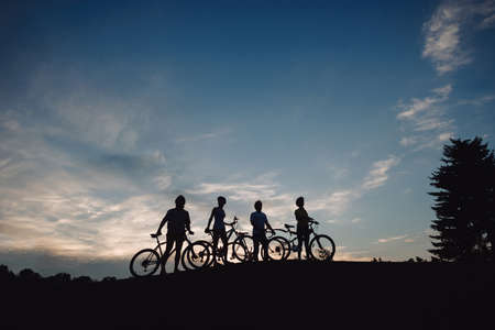 Four cyclist on hill at evening sky. Group of cyclists walking with bicycles at sunset sky. Happy...